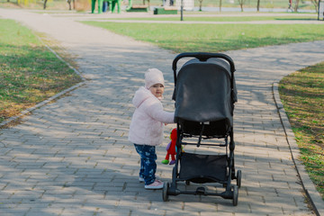 A little girl is rolling baby carriage in the park. Child in the park playing with pram.