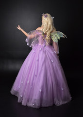  full length portrait of a blonde girl wearing a fantasy fairy inspired costume,  long purple ball gown with fairy wings,   standing pose with back to the camera on a dark studio background.