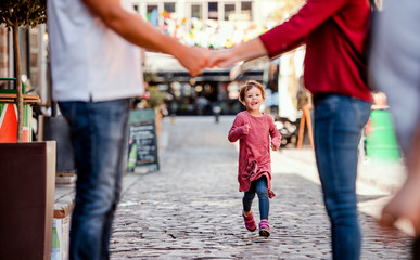 A small girl running towards unrecognizable parents outdoors in town.