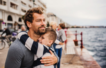 Father with small son standing outdoors in town by the sea, hugging.