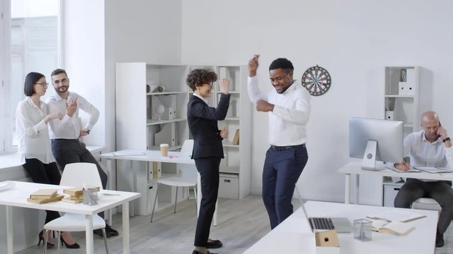Full shot of multiethnic group of coworkers having fun during break and throwing darts in pairs, with one team celebrating their win and another looking disappointed, while their boss is busy at desk