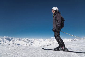Fototapeta na wymiar A bearded male skier in a helmet and a ski mask is standing on skis against the background of snow-capped mountains and a blue sky. Athlete in a black suit