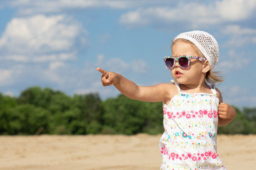 Little girl in sunglasses pointing finger  hand aside and  looking aside on summer beach