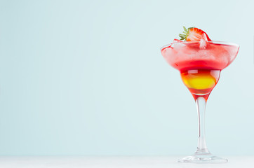 Bright layered red and yellow alcohol cocktail with strawberry slice, straw, ice in wineglass on soft light pastel blue background.