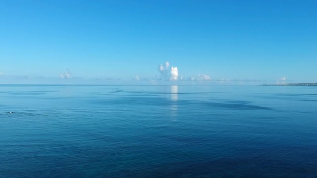 Early morning blue sky reflecting in the ocean with a few cumulonimbus clouds on the horizon. Drone dropping in altitude to give a glimpse of the reef below.
