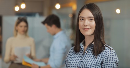 Portrait of young successful businesswoman looking at camera standing in office workspace. Working people on the blurred background.