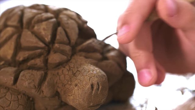 Creating toy made of clay in the workshop. Child is scratching with spicial tool the texture of the scales on the handmade turtle toy's head from clay, hand closeup.