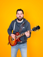 man with black hair and beard, wearing grey hoodie playing the electric guitar in front of a yellow background