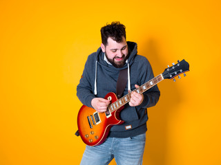 Obraz premium man with black hair and beard, wearing grey hoodie playing the electric guitar in front of a yellow background
