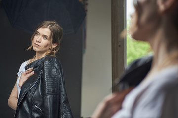 young, sympathetic, emotional, sexy, European light girl with loose hair in a black leather jacket and in a white dress near a mirror, visible reflection in the mirror. On the gray background