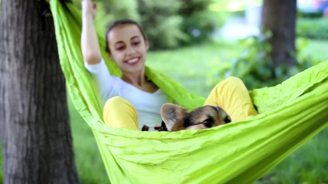 attractive woman swinging on hammock with dog. young pretty smiling woman with cute Welsh Corgi dog lying in bright green hammock in park.