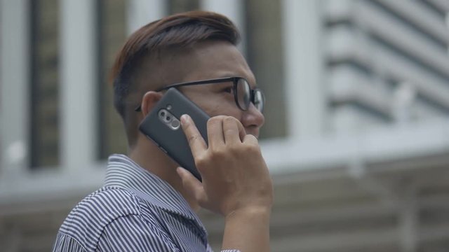 Slow motion portrait asian businessman answers the phone. Asian man with glasses use phone in street near big office building.