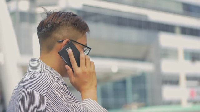 Slow motion portrait asian businessman answers the phone. Asian man with glasses use phone in street near big office building.
