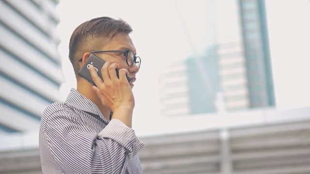 Slow motion portrait asian businessman answers the phone. Asian man with glasses use phone in street near big office building.