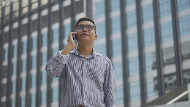 Slow motion portrait asian businessman answers the phone. Asian man with glasses use phone in street near big office building.