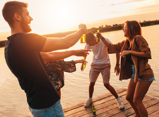 Group of young friends having fun drinking beer and dancing on pier by the lake at sunset.