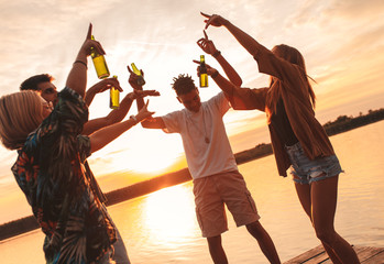 Group of young friends having fun drinking beer and dancing on pier by the lake at sunset.