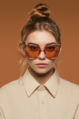 Cropped front view shot of blonde lady, wearing shirt. The girl with bun and wavy hair locks in butterfly sunglasses with light brown rim and lenses, is looking at camera against brown background.