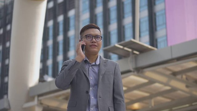 Slow motion portrait asian businessman answers the phone. Asian man with glasses use phone in street near big office building.
