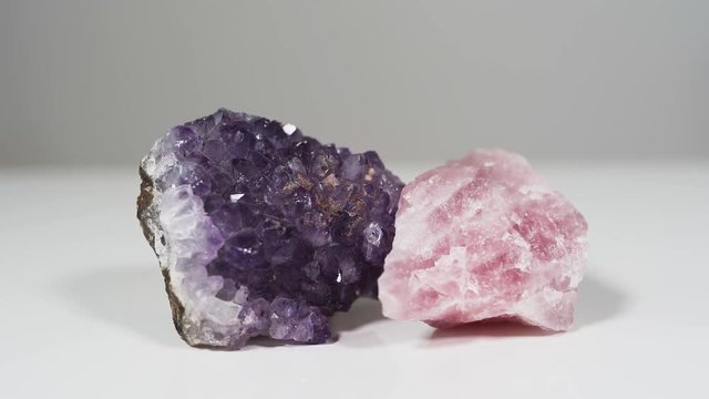 Purple Amethyst and pink Rose quartz clusters laying on a white background. The stones have different positive healing powers and are commonly used for meditation
