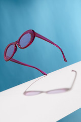 Front view shot of oval-shaped glasses with purple rim and silver glitter in lenses. The tilted accessory is isolated in the air on background with wide white stripe. Fashionable summer accessory.