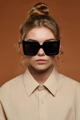 Cropped front view shot of lady, wearing shirt. The girl with bun and wavy hair locks in square sunglasses with black rim and lenses. The woman is looking at camera against light brown background.