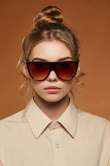 Cropped front view shot of lady, wearing shirt. The girl with bun and wavy hair locks in wrap sunglasses with ombre-brown rim and lenses. The woman is looking at camera against brown the background.