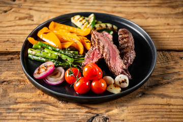 Grilled and sliced beef steak with grilled vegetables served on black plate on old rustic wood background.