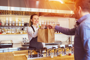 Cheerful waitress wearing apron serving customer at counter in restaurant - Small business and service concept with young woman owner offering recycled paper bag with take away food to online client - 271573933