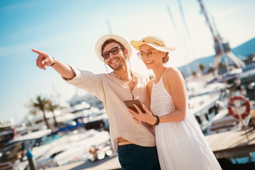 Fototapeta premium Happy young couple walking by the harbor using digital tablet of a touristic sea resort with sailboats on background