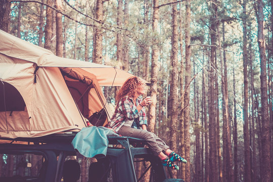 Happy free alternatie vacation traveler people - cute blonde sit down on the roof of the car with tent drinking coffee and enjoying the beauty of the forest around - wanderlust lifestyle girl