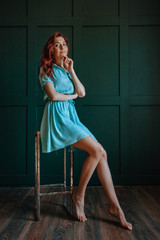 Obraz premium Redhead girl in short dress with beautiful long legs sitting on chair on dark background
