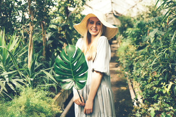 Young beautiful blonde in a dress and straw hat in a tropical greenhouse with monstera leaf in her hands, happy vacation days, summer mood concept.