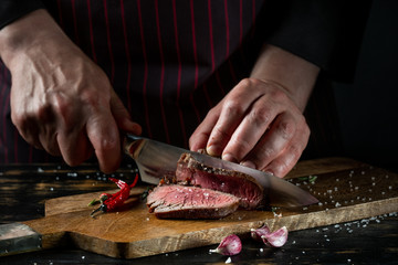 Slicing juicy beef steak by knife in chef hands closeup. Food cooking concept. Dark black background copy space. - 271580397