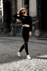 Outdoor portrait of a pretty woman walking near old buildings on a sunny day. Young female in black clothes strolls across street. Trendy girl enjoying summertime in the quaint old city
