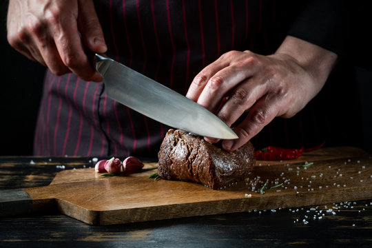 Meat steak slicing by knife in chef hands closeup. Food cooking concept. Dark black background copy space.