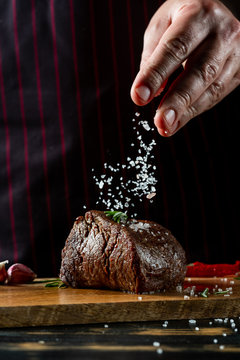 Chef hands cooking meat steak and adding salt and pepper on black copy space background for menu restaurant or recipe text.