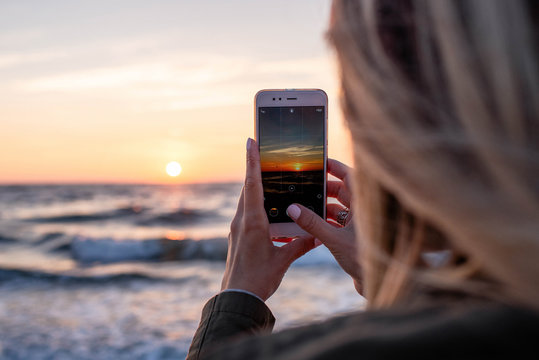 A young girl photographs the sunrise near the sea on a smartphone. A woman is holding a mobile phone. Good morning on the beach. Beautiful sky with sun and sea waves. Travel concept