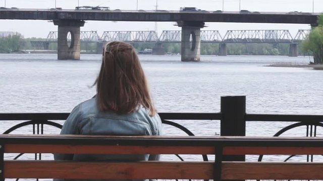 Lonely girl sitting on the bench in the park and looking on the industrial landscape