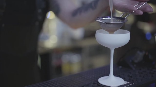 Close up hands of bartender pours liquid through stainer inside amazing white cocktail glass. Bartender mixologist combining ingredients and making tasty cocktail in a beautiful modern trendy bar.