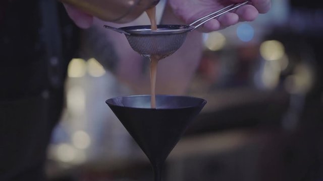 Hands of unrecognized bartender pours liquid through stainer inside cocktail glass. Bartender mixologist combining ingredients and making a tasty cocktail in a beautiful modern trendy bar.