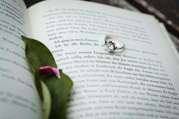 The engagement ring lies on the pages of an open book. The leaves and petals of flowers lie on the spread of the book.
