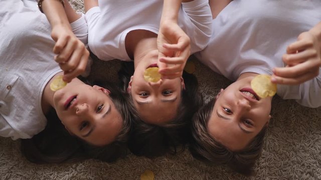 Funny eating snacks potato chips teenager triplet sisters girls lie on carpet floor top view throwing it up and catch with mouth