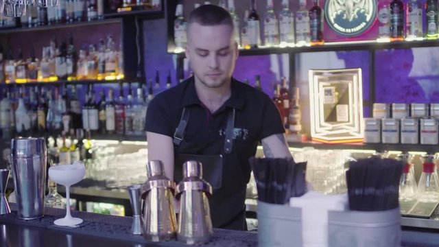 Successful professional bartender pours liquid through stainer inside amazing white cocktail glass. Barman mixologist combining ingredients and making tasty cocktail in a beautiful modern trendy bar.