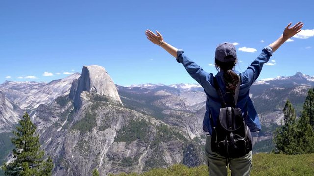 back view of cheering asian woman backpacker traveler open arms standing on mountain peak in half dome trail in yosemite. young girl hiker climbing in nature. carefree female relax outstretched hands