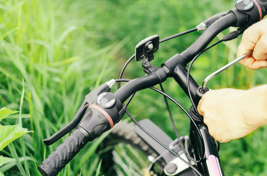 A man unscrews the bolts with a hex wrench on the handlebars of a mountain bike against the background of grass. Repair in the forest road. Close-up