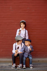 Cute children, boy brothers in vintage cloths, eating lollipop ice cream, sitting on vintage suitcase in front of a red brick wall
