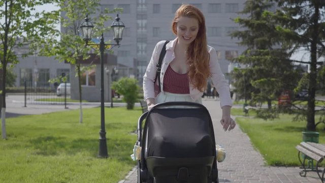 Young happy redhead mother walking with baby carriage and smiling along the street on a nice spring day