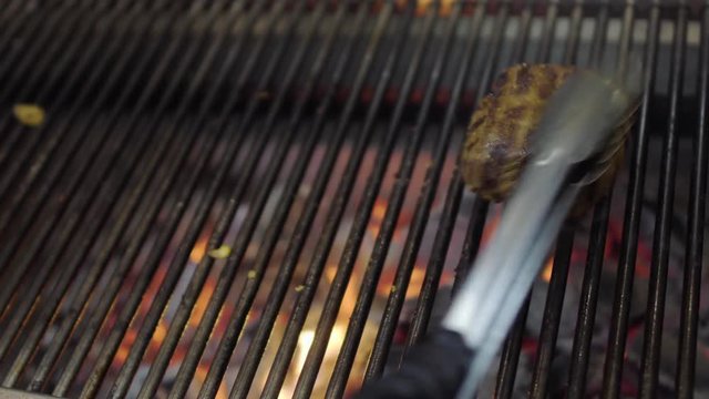 Chef preparing tasty meat on the grill, turning it with tongs close-up. Red charcoals on the bottom. Food preparation in the restaurant kitchen