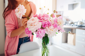 Woman puts peonies flowers in vase. Housewife taking care of coziness and decor on kitchen. Composing bouquet.